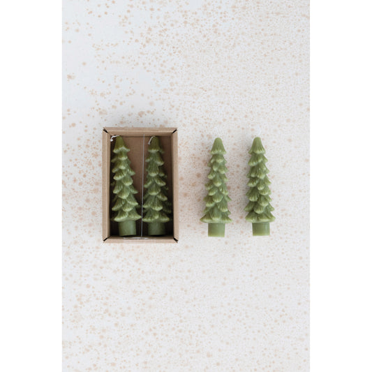 Two green tree-shaped candles on a textured white surface with one candle outside its box.