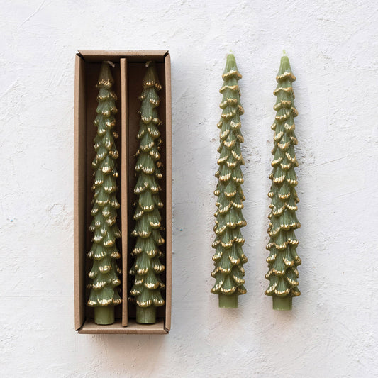 Two green tree-shaped candles with gold details on a white surface, one in an open box.