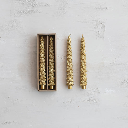 Two gold tree shaped candles on a textured white surface with one candle out of the box.