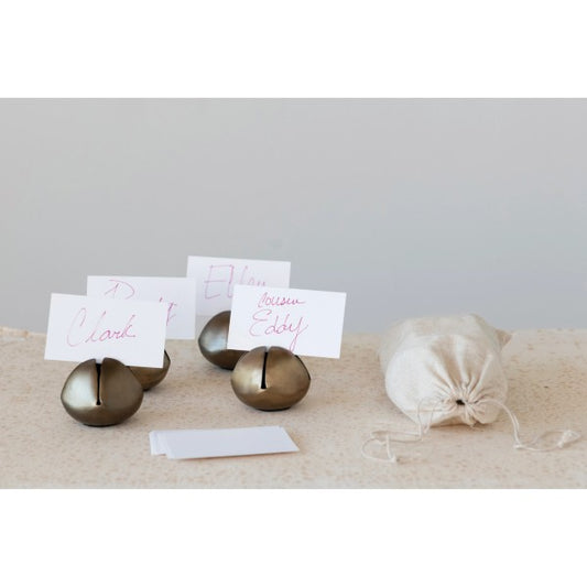 Four round metal bell-shaped place card holders with cards displaying names, placed on a surface with a drawstring bag beside them.