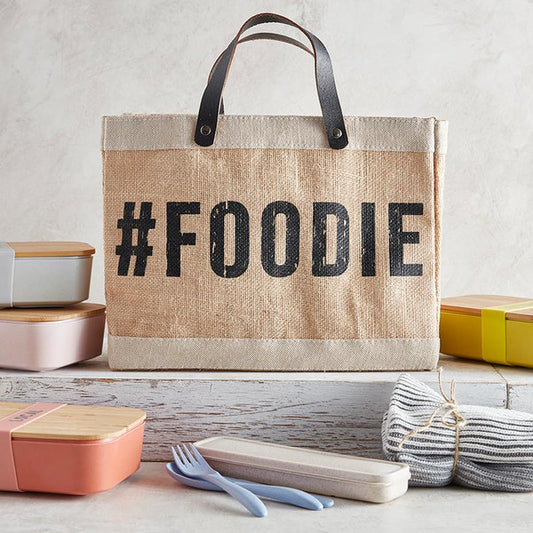 Jute bag with '#FOODIE' text on a table with food containers and utensils.