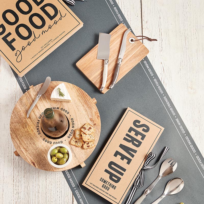 Wooden cutting board with food items and serving utensils on a gray surface with 'Good Food' and 'Serve Up' signs.