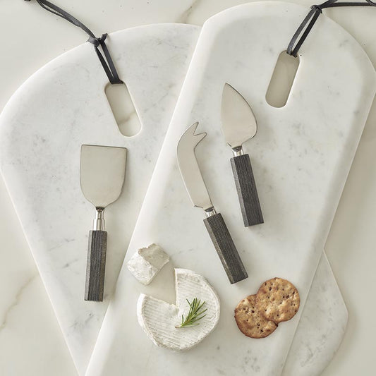 Set of cheese knives with wooden handles on a marble surface with a small dish of cheese and crackers.